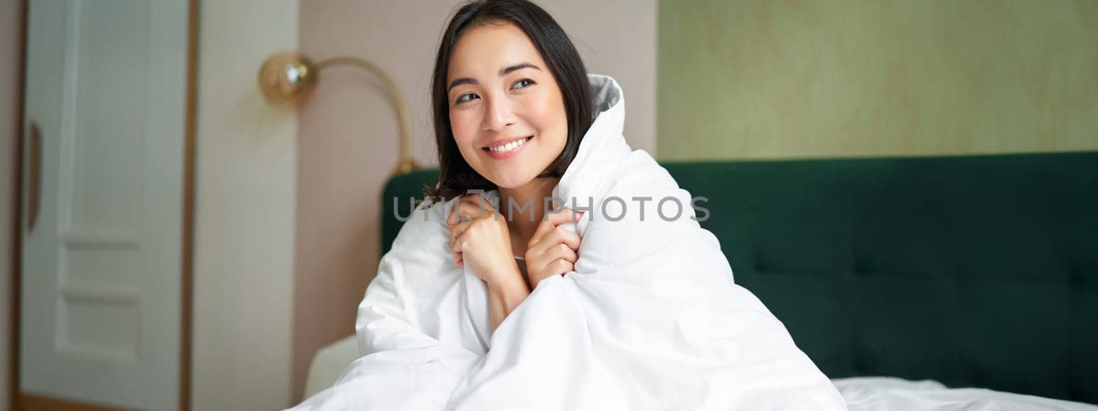 Beautiful romantic woman covers herself with warm duvet, enjoys sleeping and staying in bed, waking up happy in morning, sitting in bedroom by Benzoix