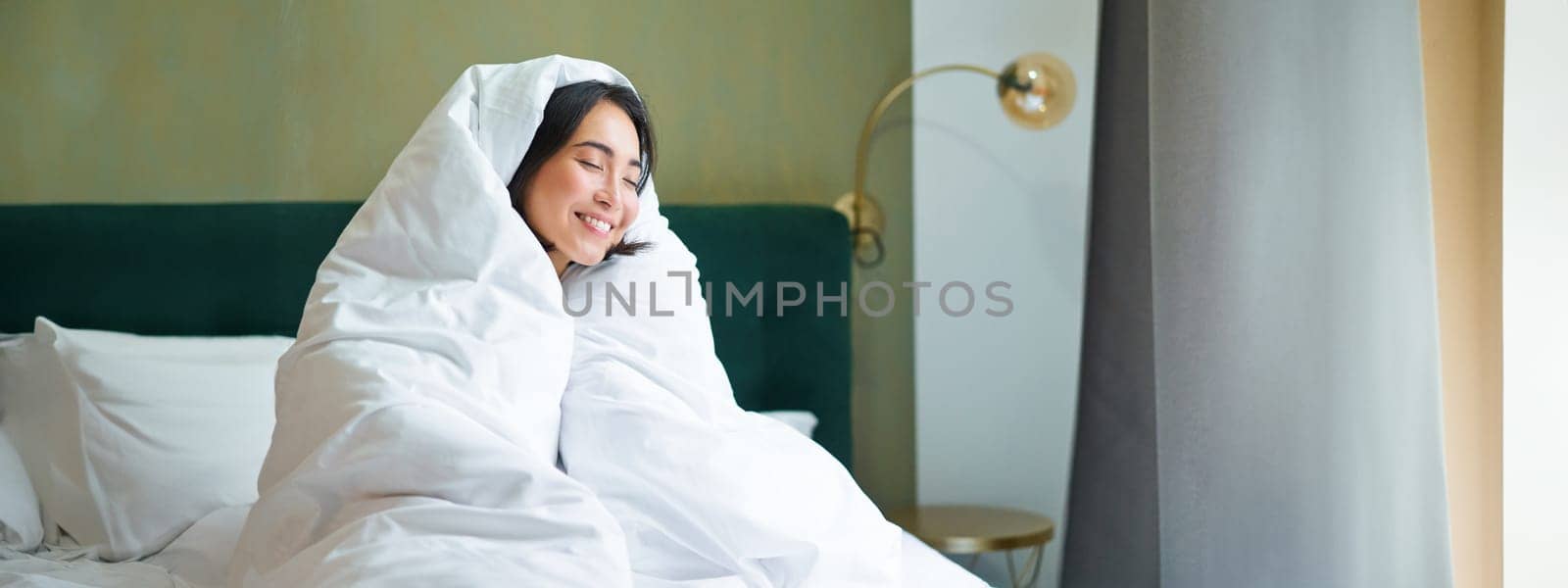 Cozy mornings. Happy asian girl feels warm in her bed, covers herself with cozy duvet in her bedroom, a comfortable stay in hotel room.