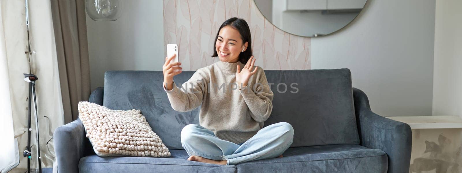 Portrait of asian woman sitting on sofa with smartphone, waving at mobile phone screen and waving at camera, video chat, talking to someone by Benzoix