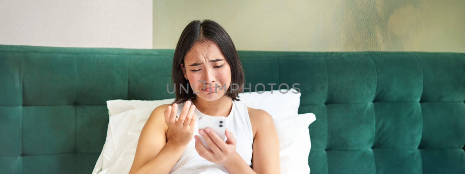 Portrait of sad frowning korean girl, lying in bed and looking at smartphone with upset, disappointed face expression, using mobile phone, complaining.