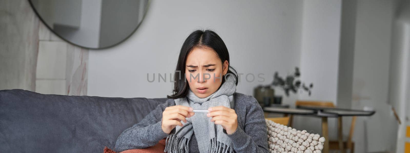 Shocked woman looks concerned at her thermometer, catching cold, has fever, sitting on sick leave at home.