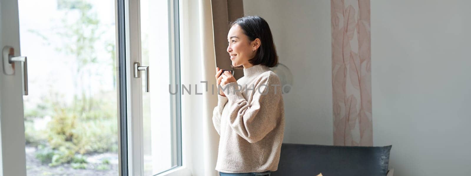 Beautiful korean woman staying at home, looking outside window, drinking coffee espresso and smiling, feeling comfort and warmth.