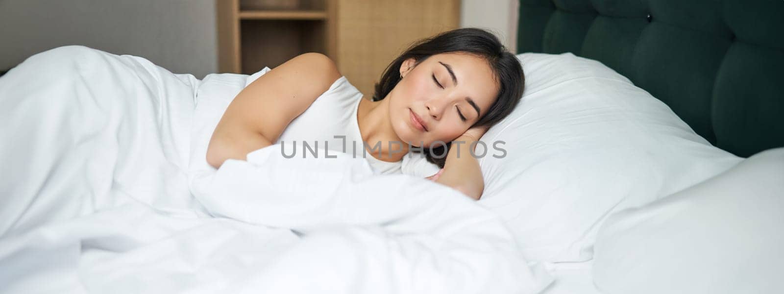 Beautiful asian girl sleeps in her bed with white sheets, warm winter duvet, smiling in her sleep, dreaming, lying in bedroom in morning.