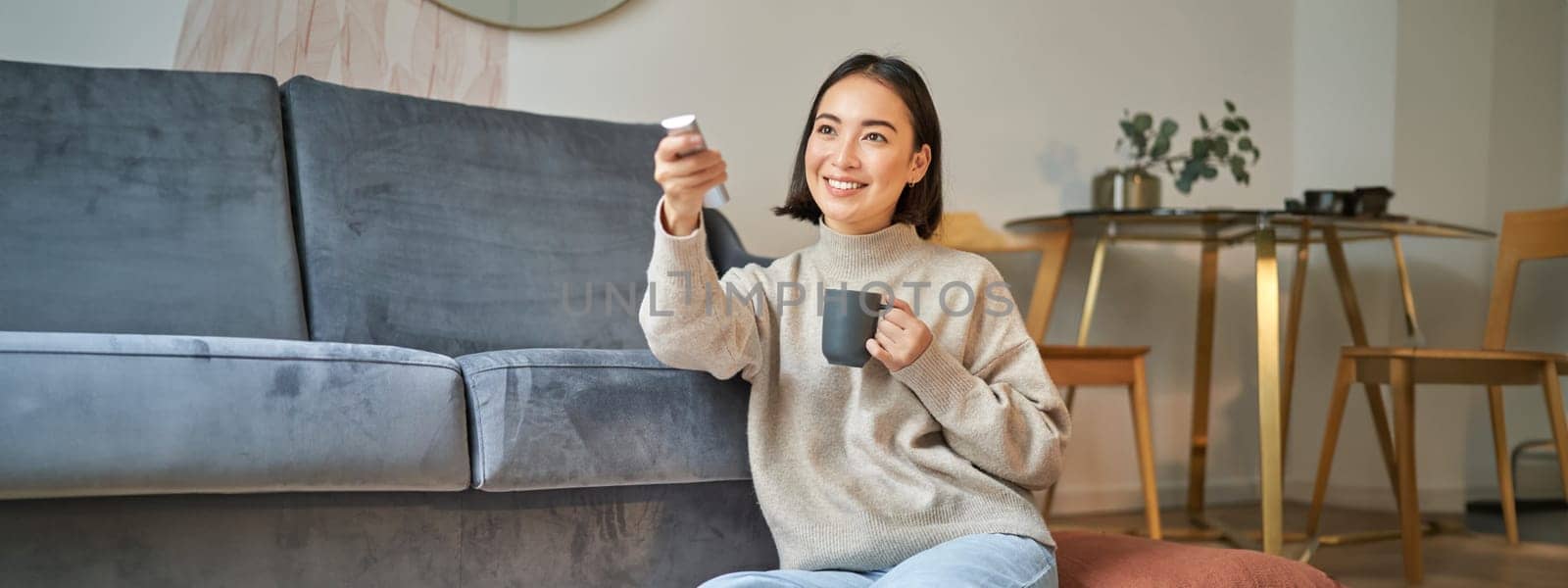 Portrait of smiling korean woman sitting near tv, holding remote and switching channels while drinking hot coffee.