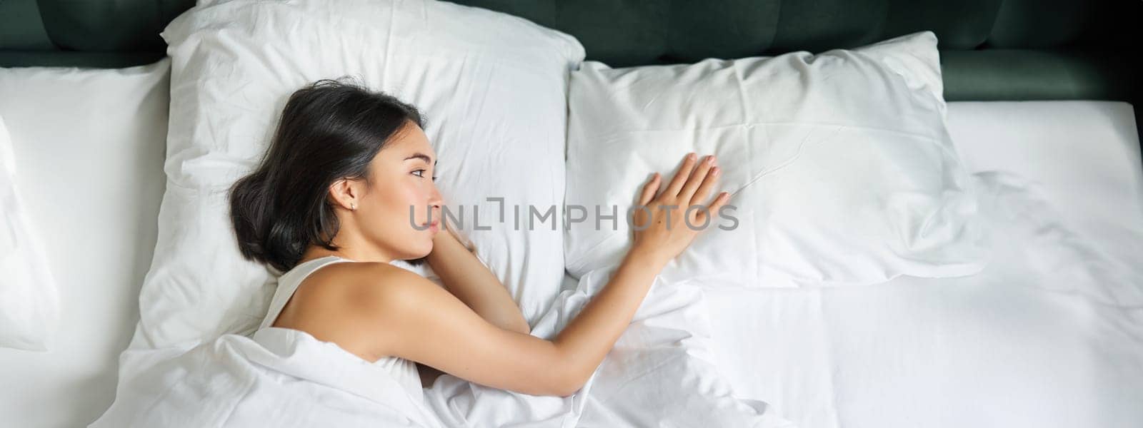 Portrait of romantic asian woman lying alone in her bed, touching empty pillow and thinking about her partner, breakup relatioship.