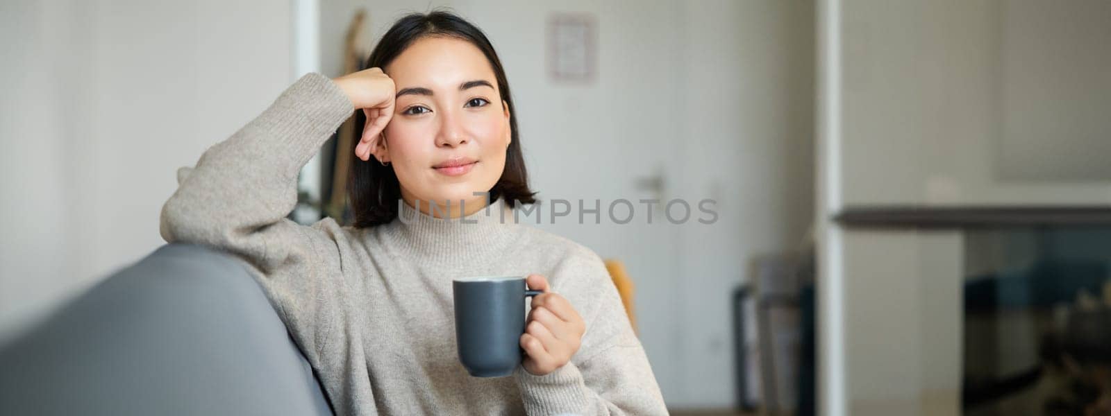 Smiling asian woman sitting on sofa with her mug, drinking coffee at home and relaxing after work, looking calm and cozy.