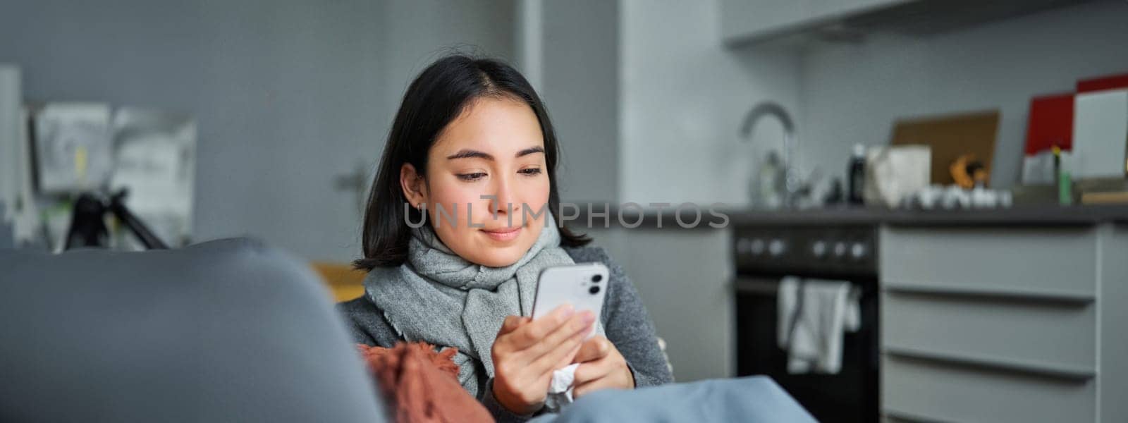 Portrait of ill young korean woman sitting on sofa, texting message, using mobile phone to contact her doctor while being sick.