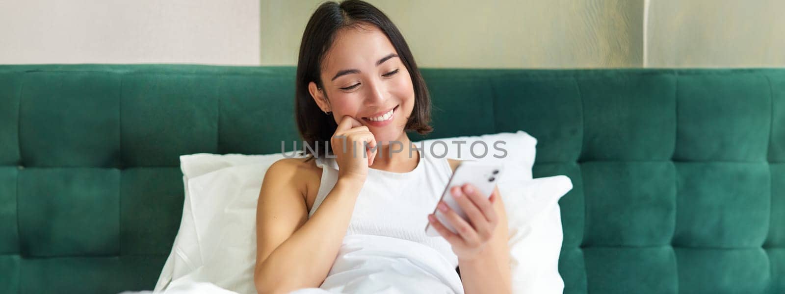 Beautiful korean woman reading mobile phone, messaging or watching video while relaxing in her bedroom, holding smartphone by Benzoix