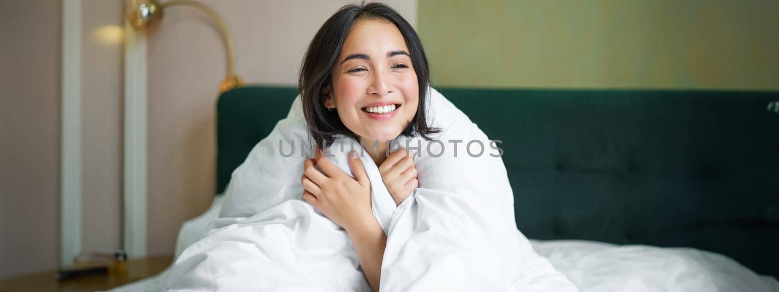 Cute and tender woman sitting in bed with warm duvet, smiling as looking out of the window, enjoying morning and comfort of blanket.