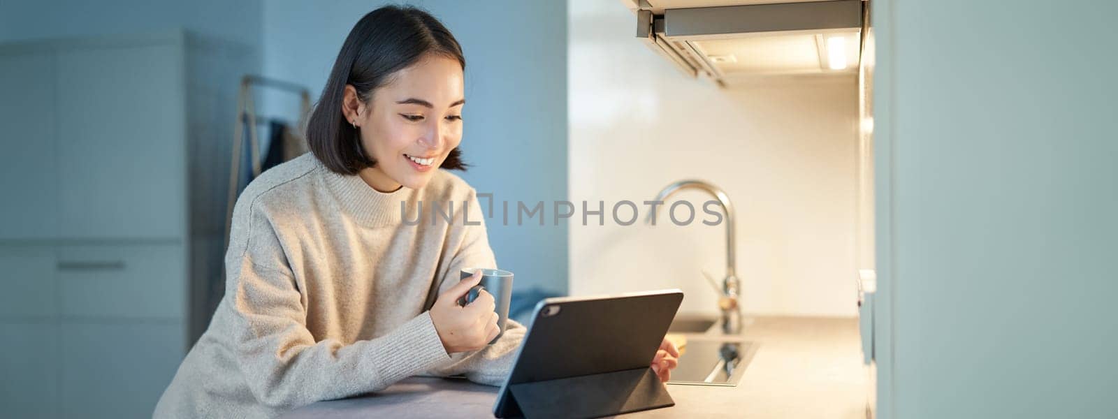 Portrait of stylish young asian woman watching videos on tablet, sitting in kitchen and drinking coffee.