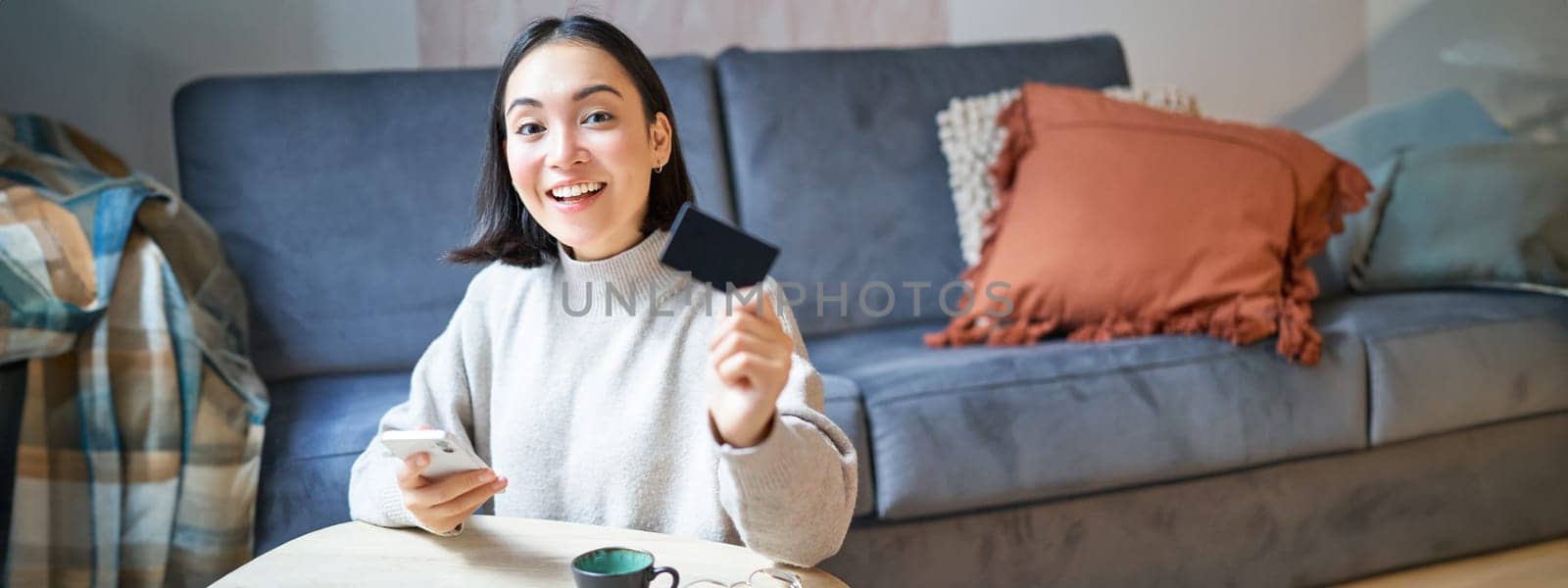 Young woman in living room, showing her credit card and using smartphone to pay, order online or shopping on application.