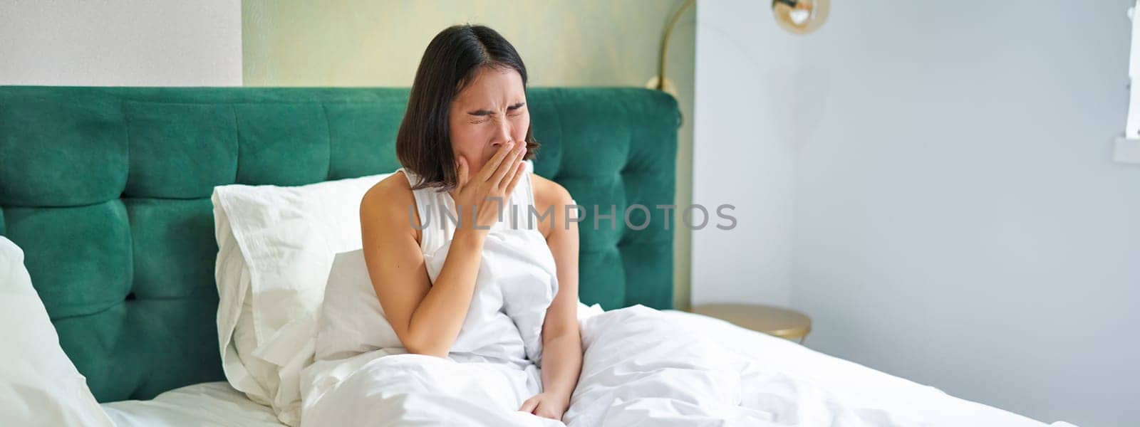 Woman waking up in bed, yawning and grimacing, early morning after sleep, sitting in bedroom.