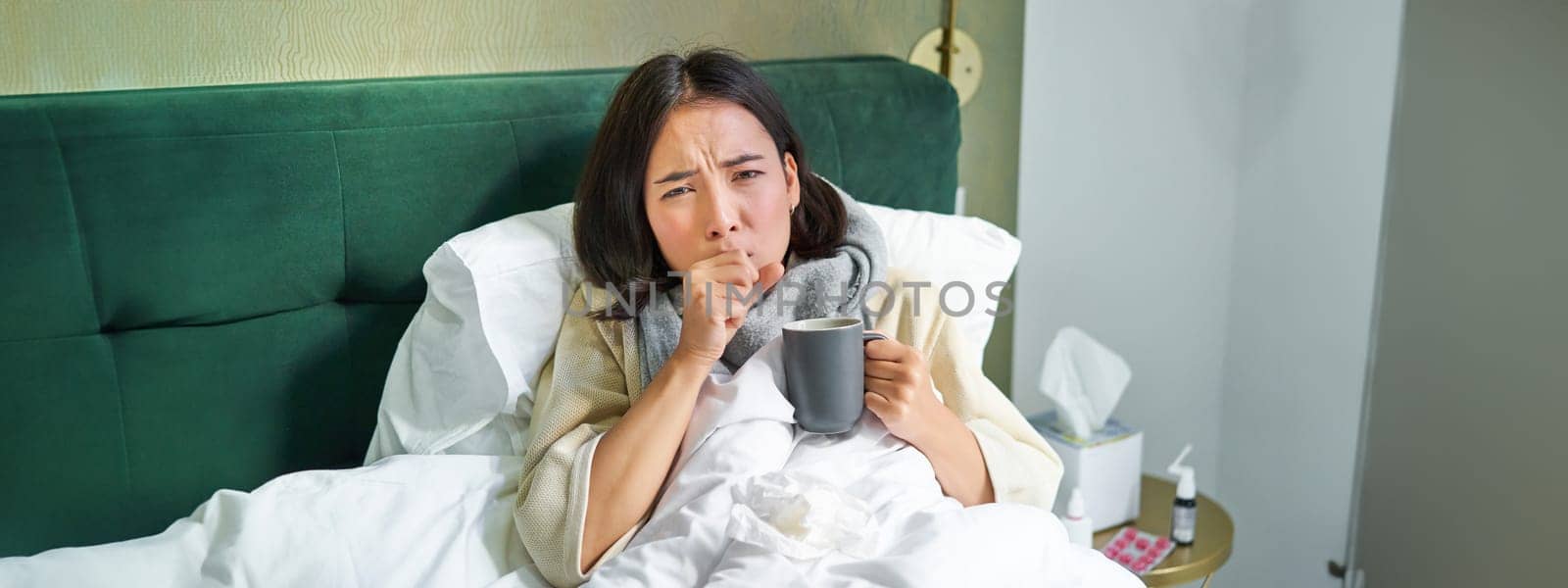 Portrait of couching asian girl in bed, feeling sick, catching cold and staying at home, looking unhealthy, drinking hot tea.