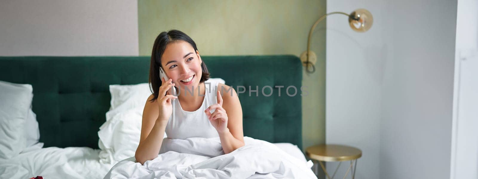 Beautiful young korean woman calling, having a phone call in bed, talking with friend while staying in bedroom by Benzoix