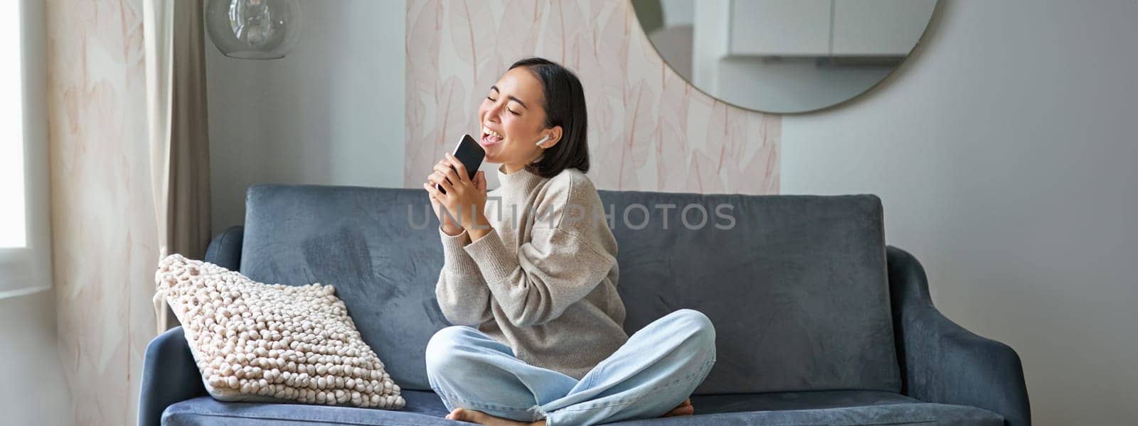 Portrait of carefree asian girl sits at home, listens to music in headphones and sings at smartphone, holds mobile phone like microphone.