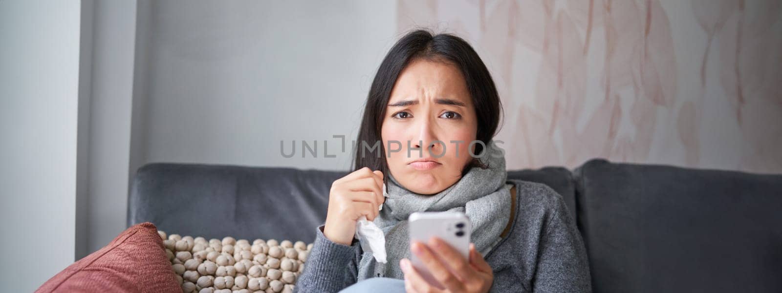 Asian girl with influenza, sits at home, cries and looks upset, holds smartphone, feels unwell and gloomy, wearing warm clothes by Benzoix