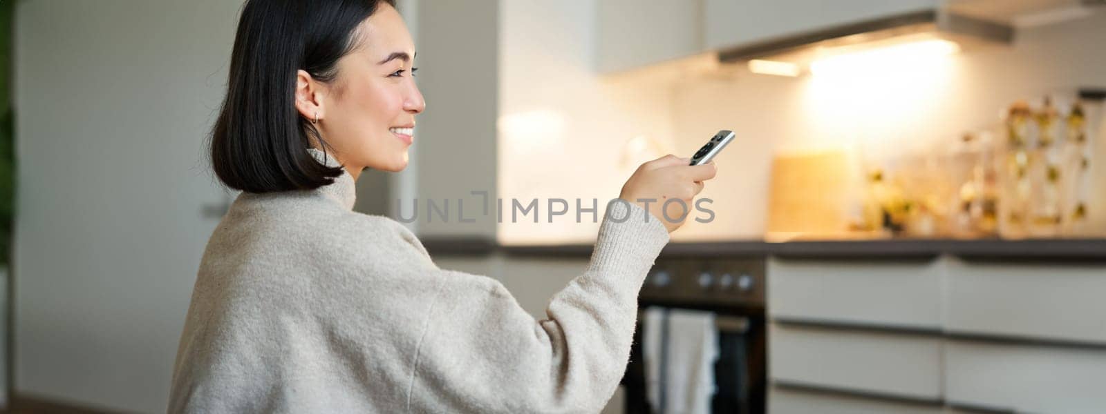 Close up portrait of smiling asian woman watching television, sitting on sofa with remote and switching chanel, looking relaxed.