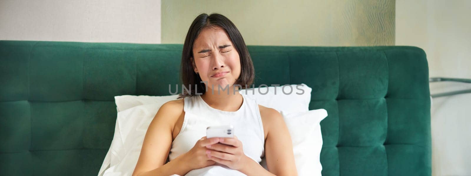 Disappointed asian girl, crying and whining, feeling upset, lying in bed on pillow, holding mobile phone, read upsetting bad news, waking up in awful mood.