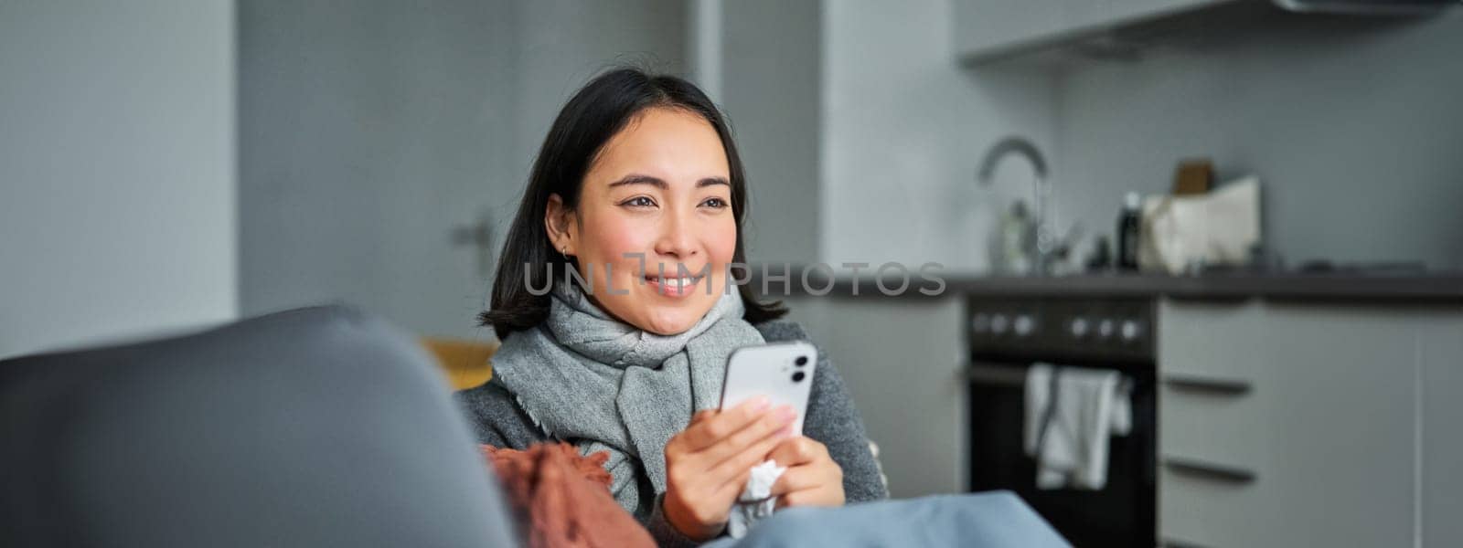 Portrait of ill young korean woman sitting on sofa, texting message, using mobile phone to contact her doctor while being sick.