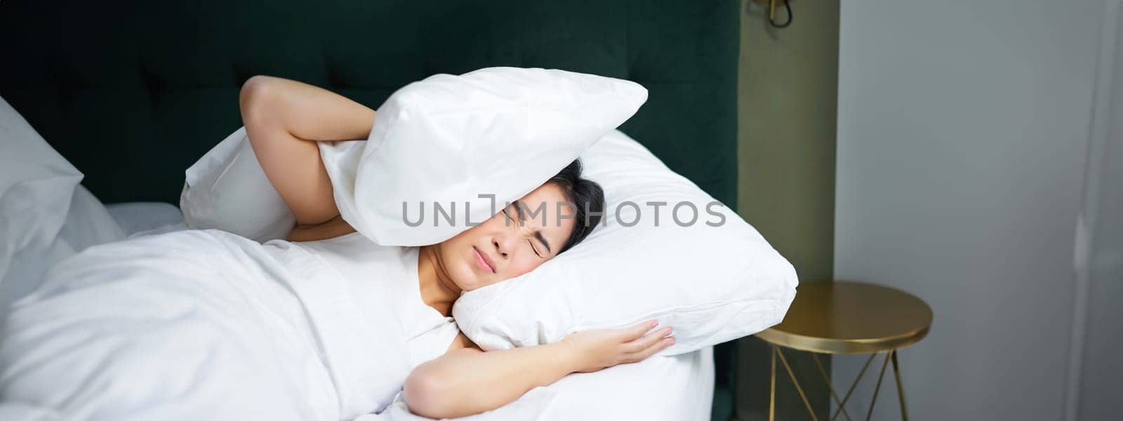 Insomnia. Asian girl shuts her ears with pillow as partner snoring. Woman lying in bed, bothered by noise at night, cannot sleep.