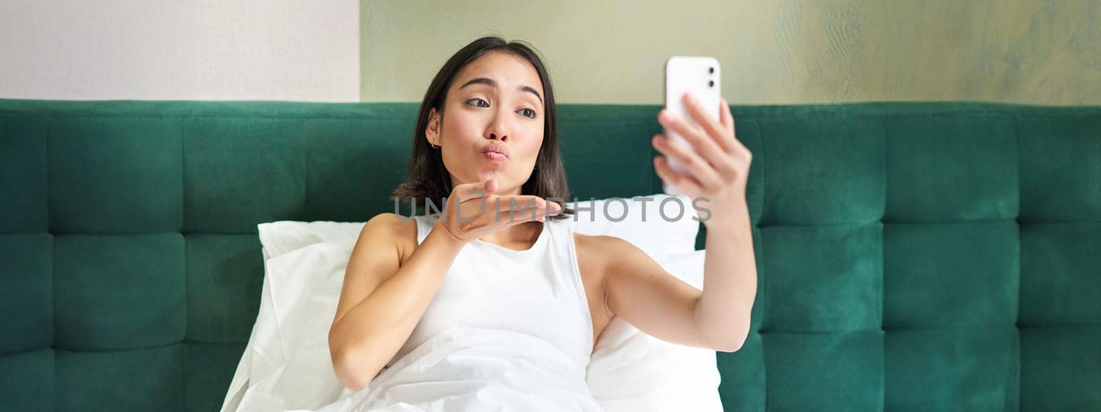 Beautiful asian girl lying in bed, making morning selfie, taking picture on smartphone in bedroom, smiling happily.