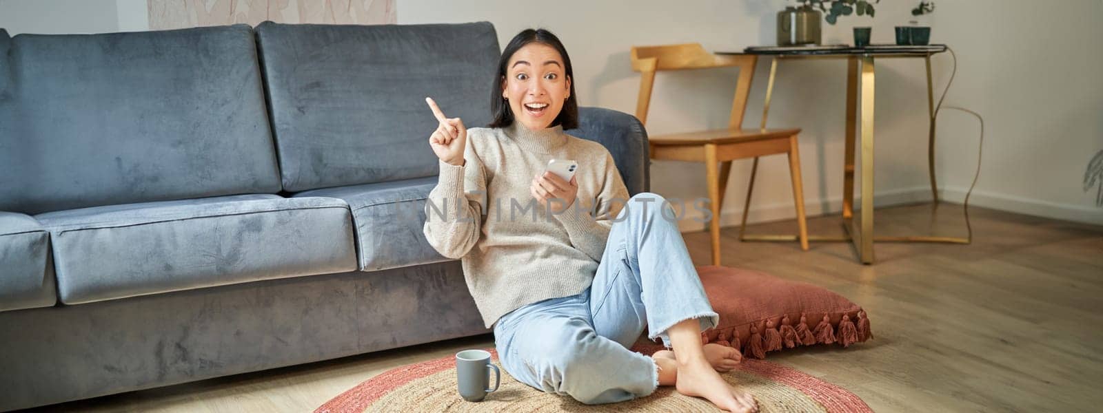 Smiling asian girl sits on floor in stylish living room, pointing finger at advertisement, showing promo banner, holding mobile phone in hand by Benzoix