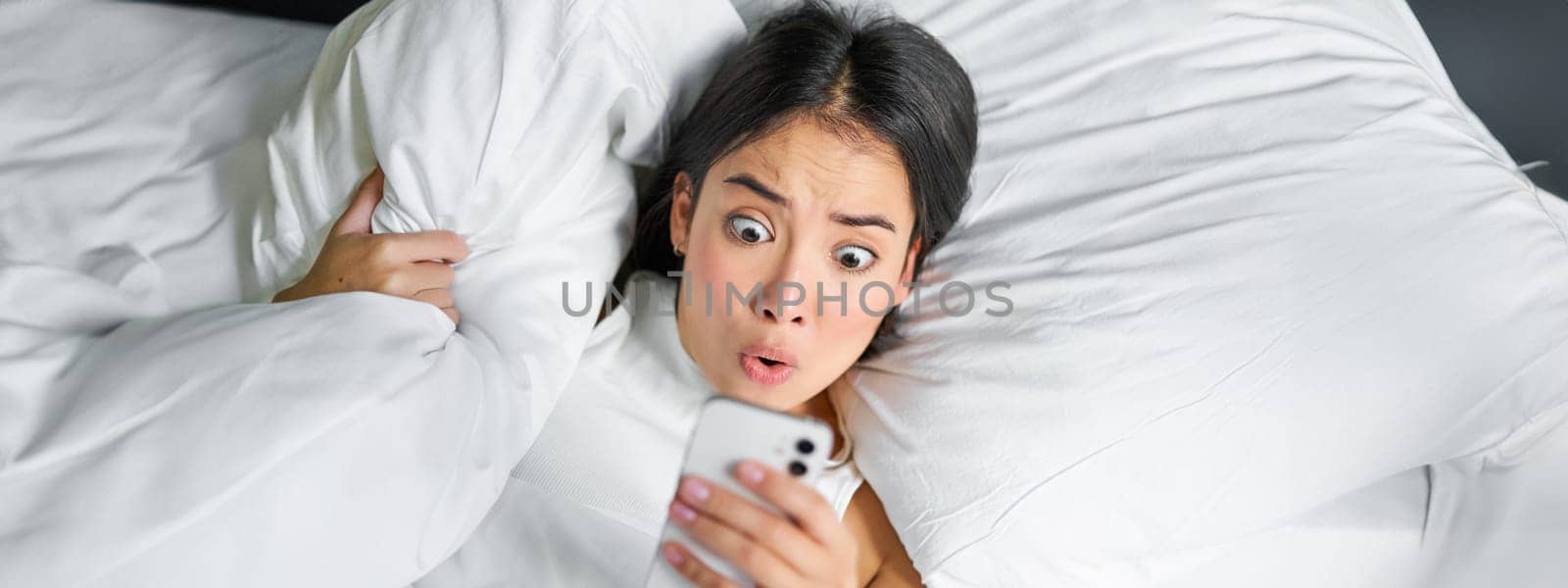 Portrait of asian girl wakes up in morning, looks at mobile phone with shocked face, overslept, hugging pillow and grimacing.