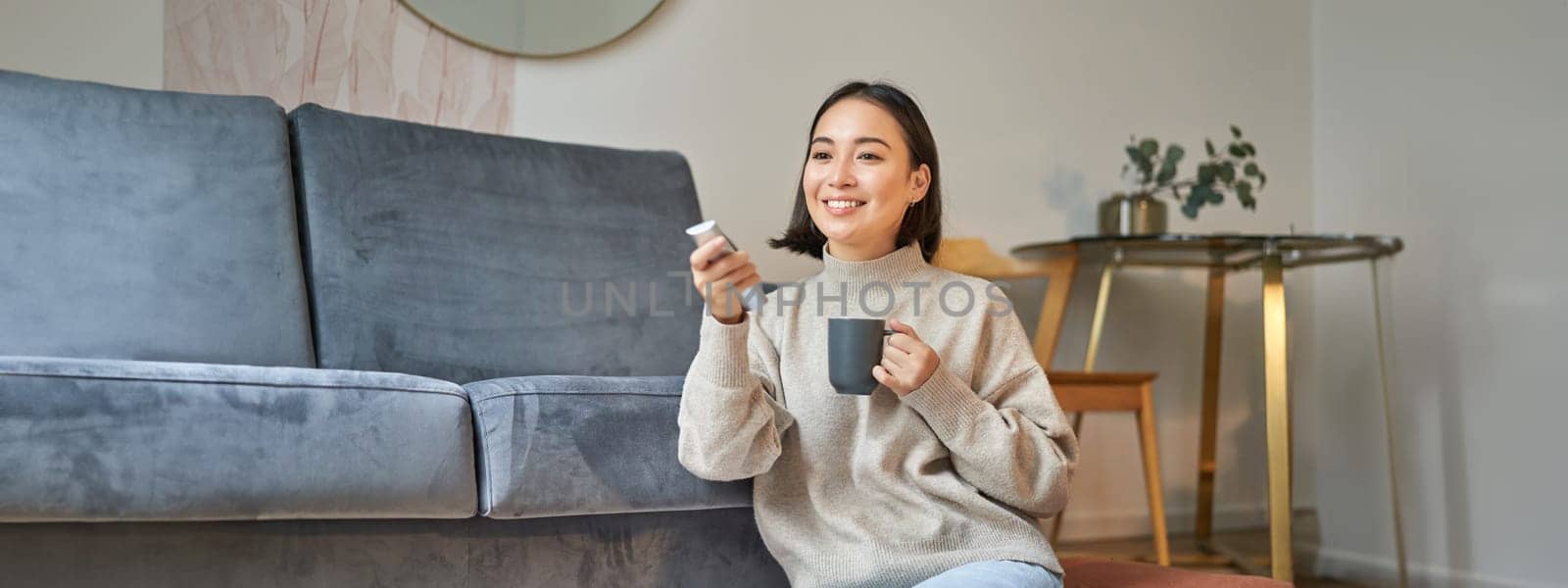 Portrait of smiling korean woman sitting near tv, holding remote and switching channels while drinking hot coffee.