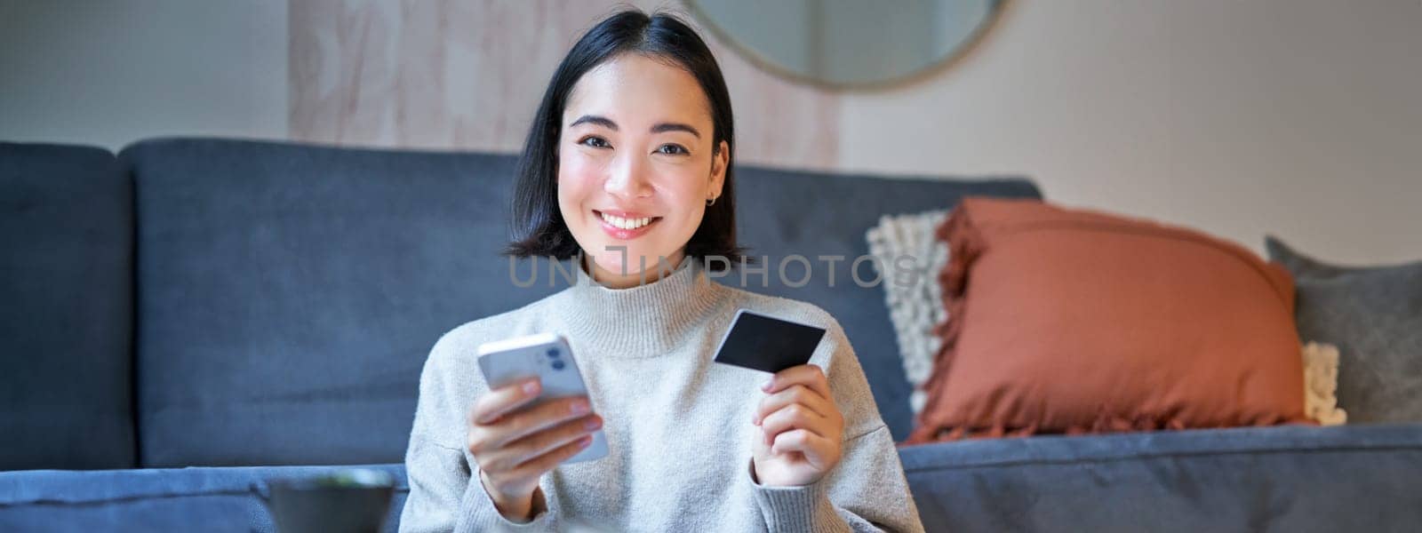 Portrait of smiling korean woman using credit card and smartphone app, paying for purchase, order online delivery.