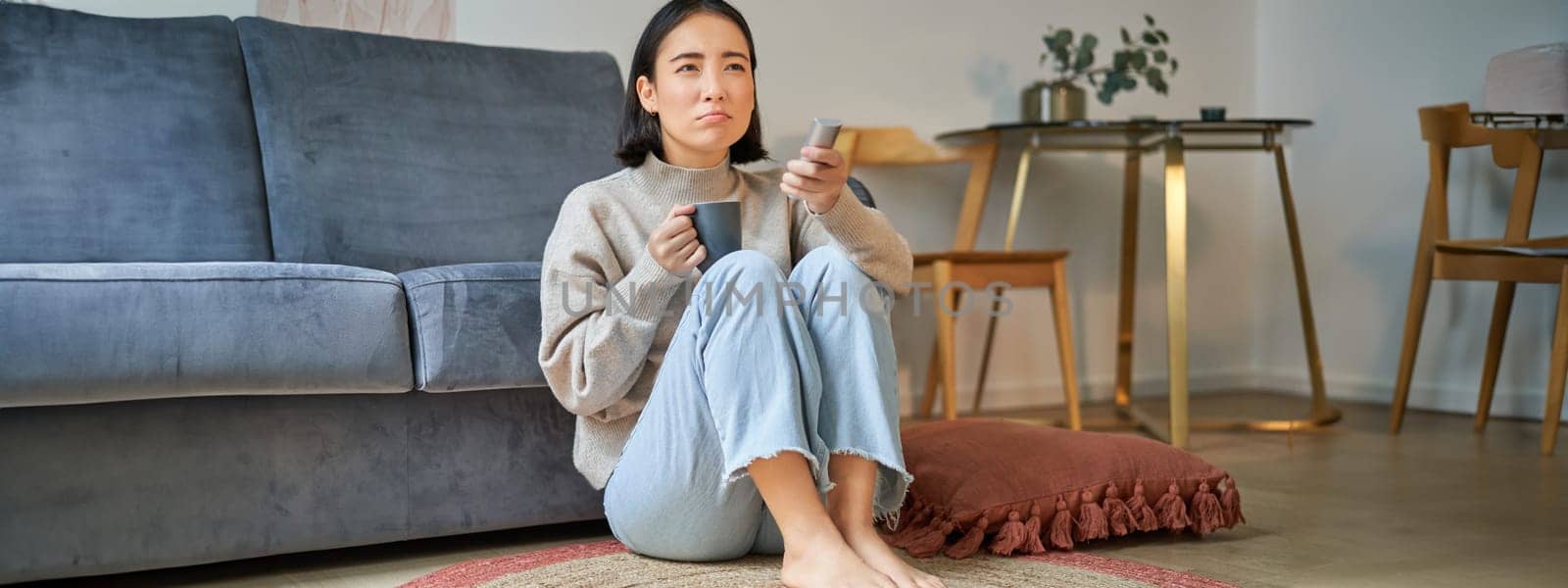 Image of young asian woman with remote, watching tv, drinking coffee, looking suspicious at television screen.