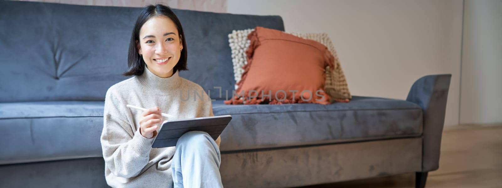 Vertical shot of cute asian girl in cozy home, drawing on her digital tablet with pen, designer working on project by Benzoix