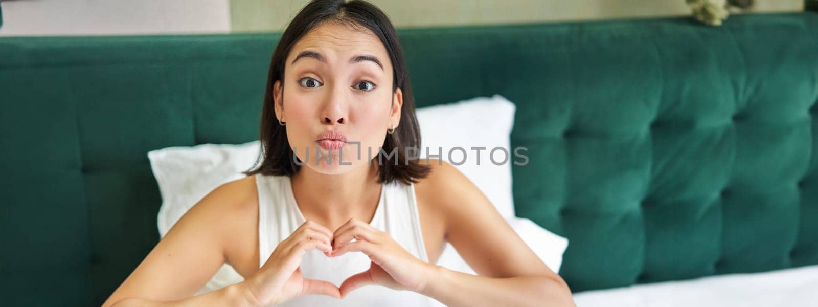 Cute asian girl shows heart, love sign, lying in bed and smiling, enjoying mornings, lazy days in bedroom by Benzoix