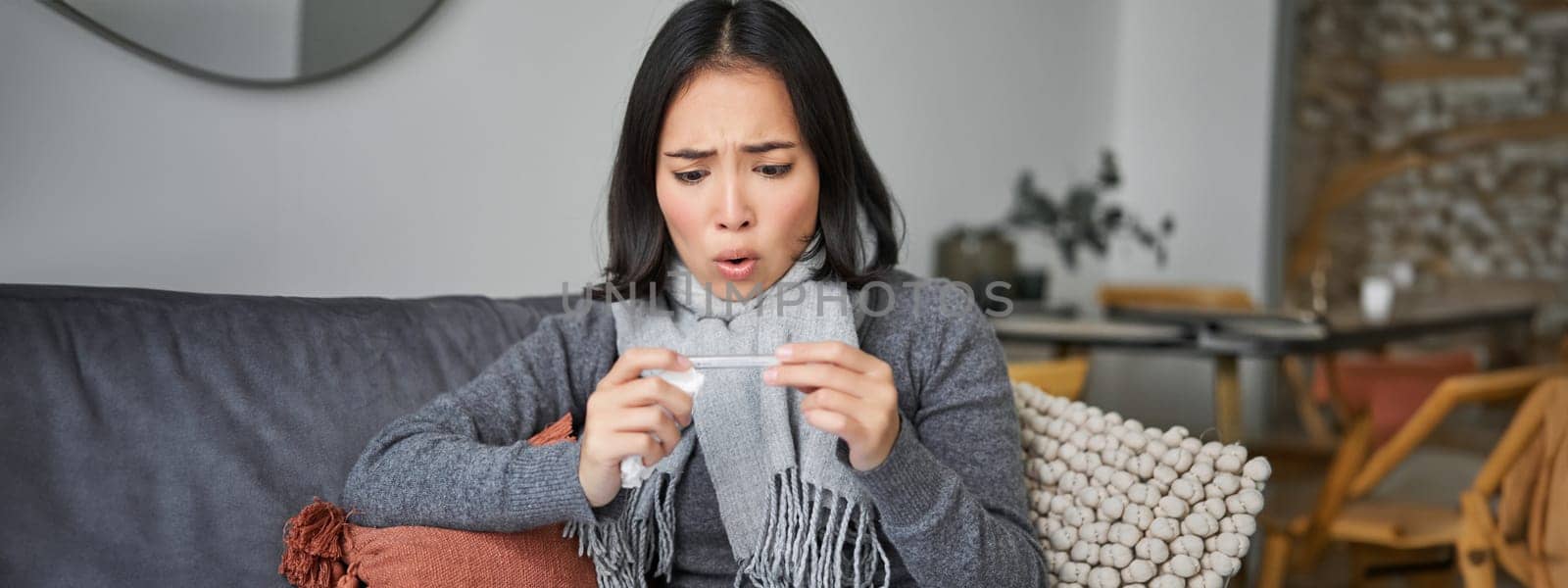 Shocked woman looks concerned at her thermometer, catching cold, has fever, sitting on sick leave at home.