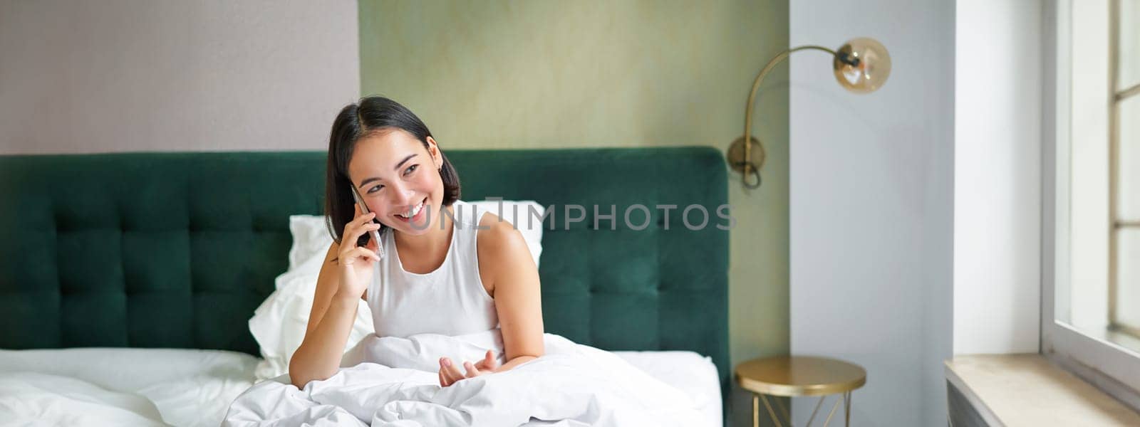 Smiling korean girl talks on mobile phone and lying in bed. Cute woman answers telephone call, holds smartphone, relaxing in her bed by Benzoix