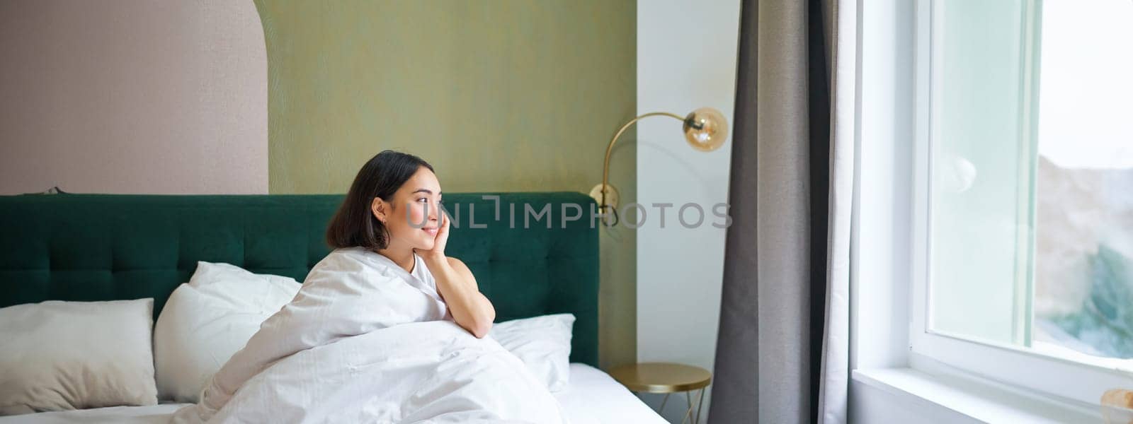 Beautiful sleepy asian girl wakes up in her bed with cozy white blankets, looking outside window with pleased smile. Lifestyle concept.