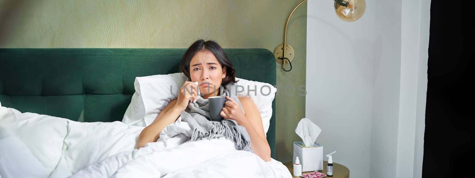 Health and people. Asian girl lying in bed, feeling sick, sneezing and drinking hot tea, catching cold, staying at home on leave.