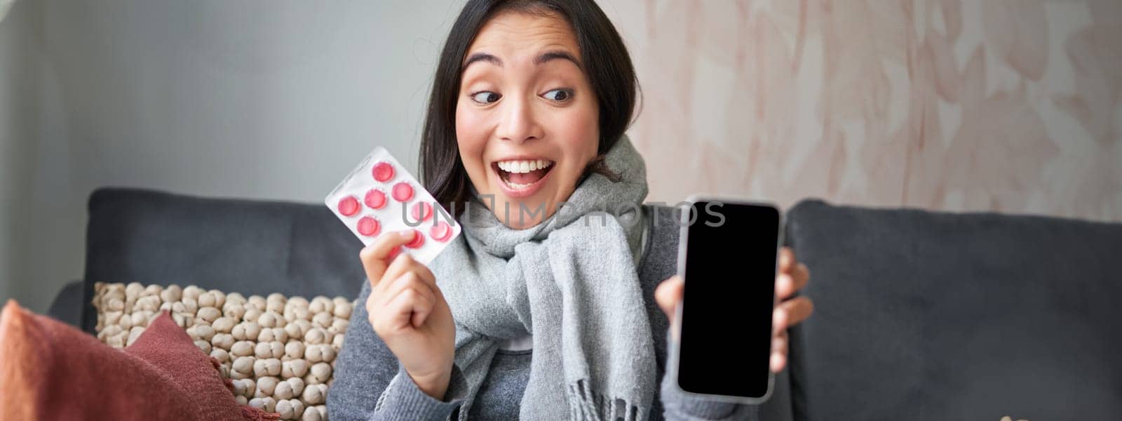 Happy asian woman catching cold, showing smartphone screen and pills, recommending online doctor app, GP or medical clinic on phone, holding drugs, staying at home with flu,