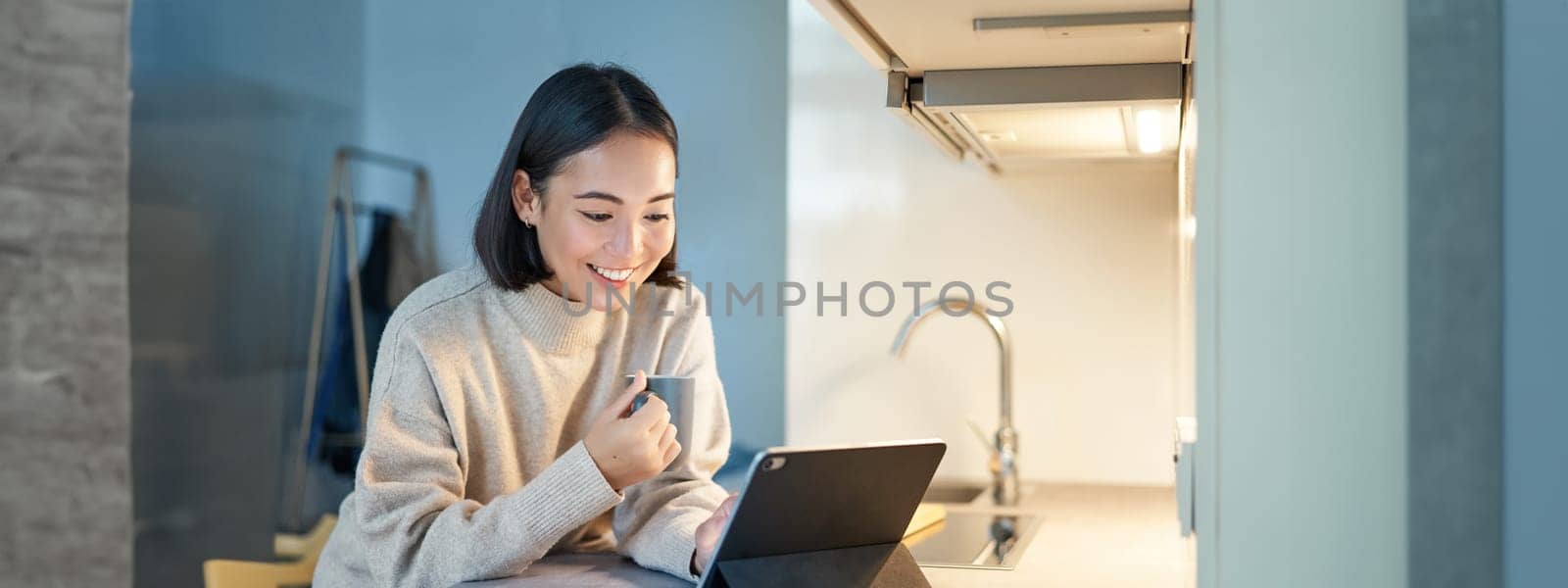 Portrait of stylish young asian woman watching videos on tablet, sitting in kitchen and drinking coffee by Benzoix