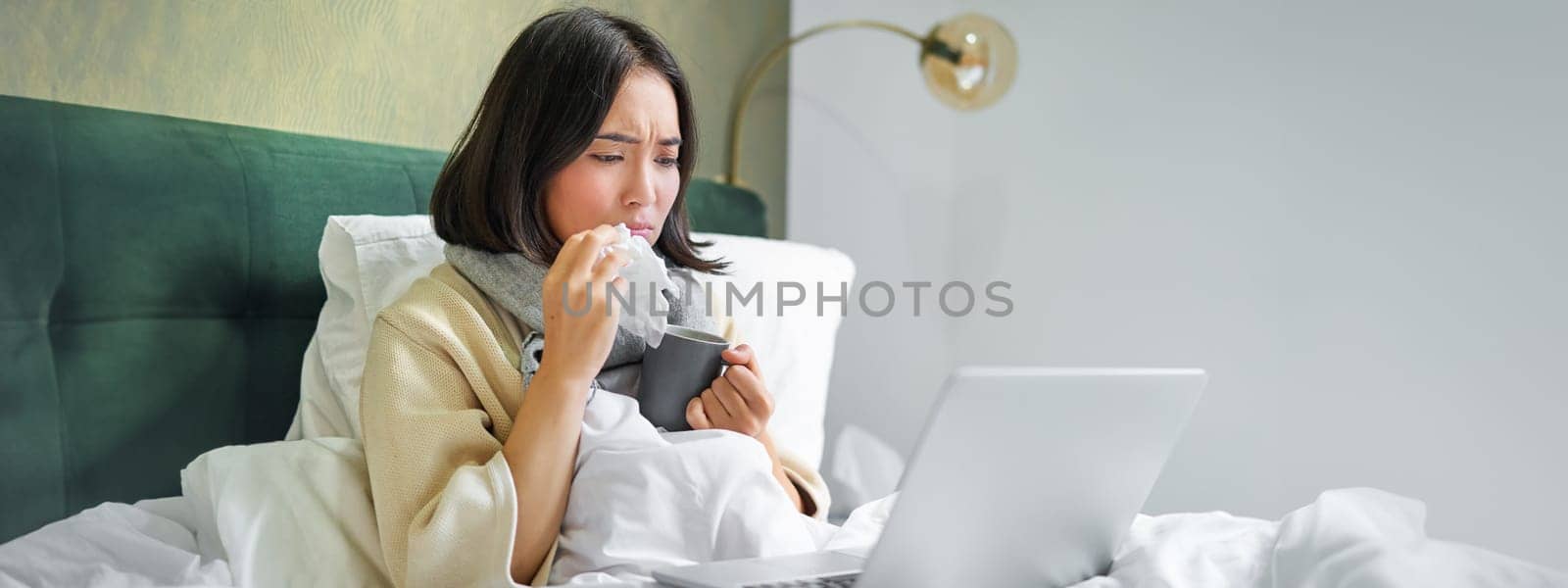 Portrait of asian girl sneezing and wiping runny nose, feeling sick, staying in bed with influenza, looking at laptop.