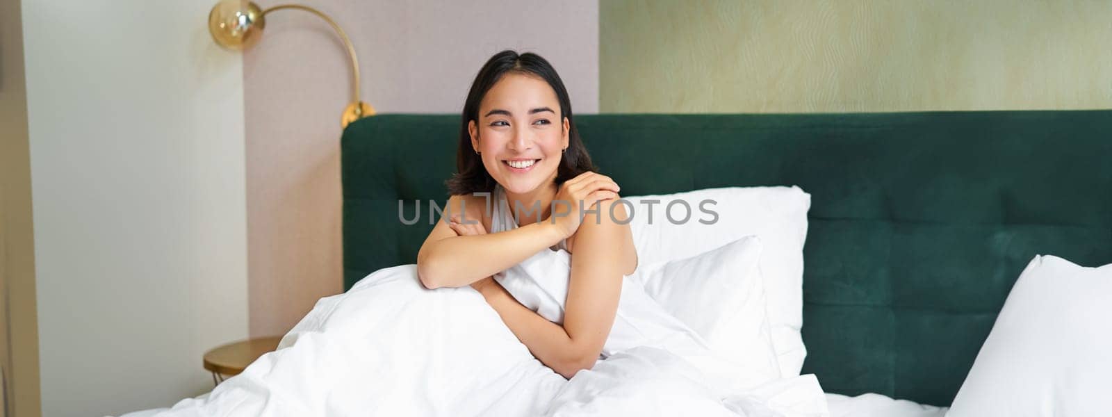 Happy mornings. Smiling asian woman wakes up in her bed, looks outside and feeld enthusiastic about her morning. Bedroom interior concept.