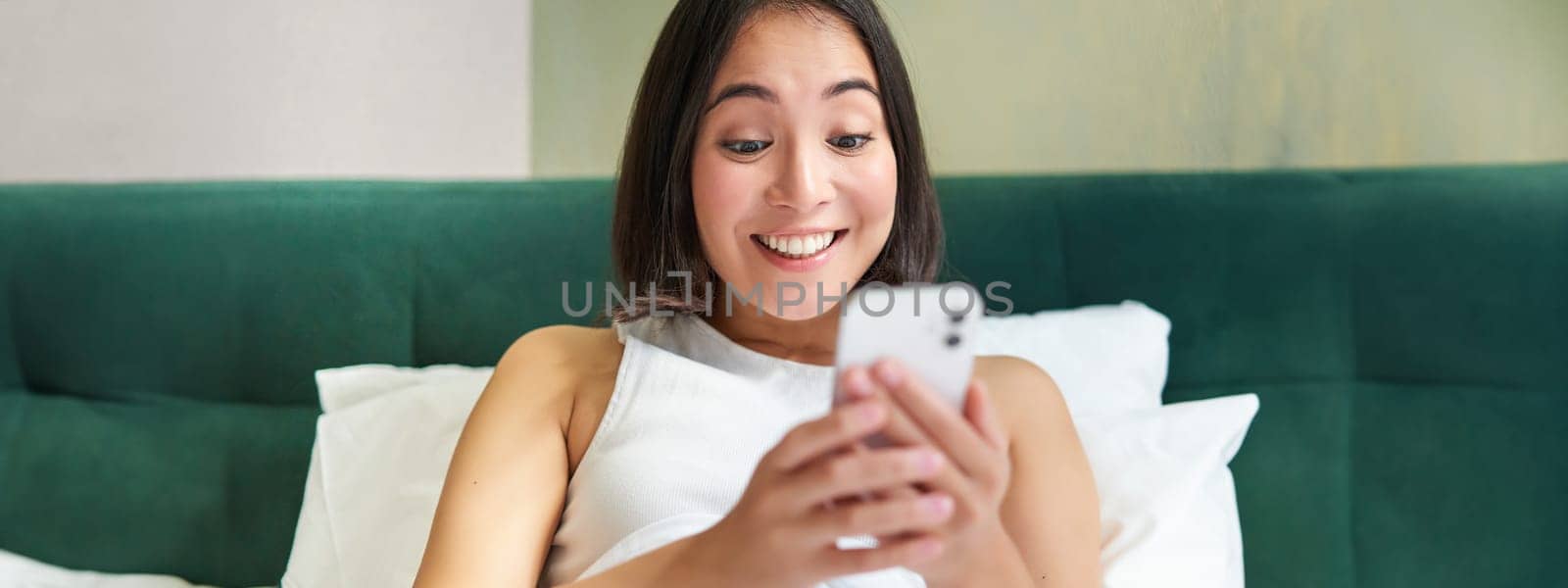 Portrait of smiling asian woman lying in bed, looking at smartphone, using mobile phone with happy face, relaxing on lazy morning.