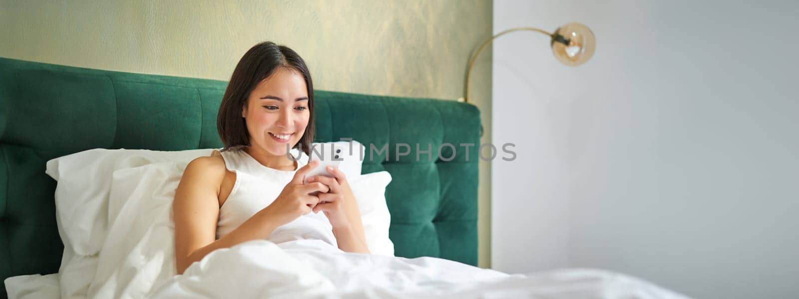 Beautiful smiling asian woman in bed, reading message on smartphone and laughing, enjoying lazy day in bedroom, using mobile phone.