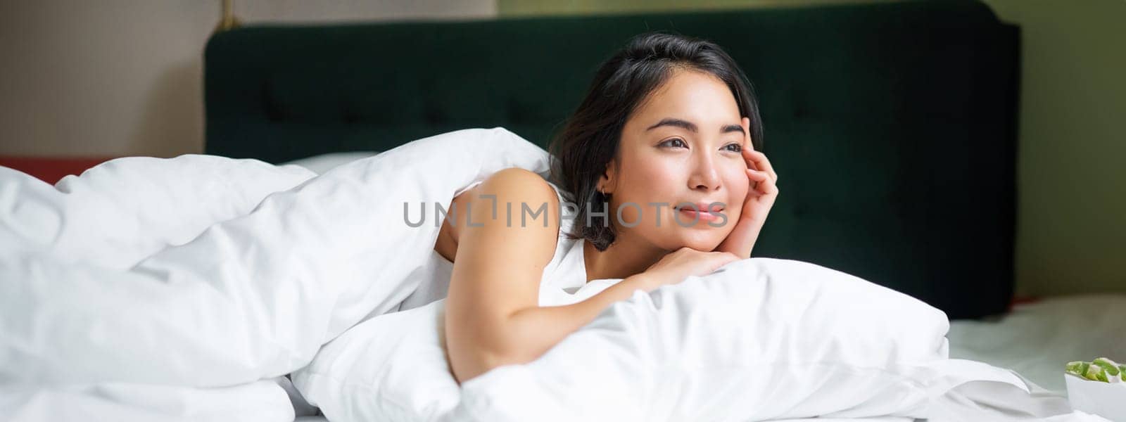 Dreamy asian girl lying in her bed in morning, covered with warm duver and leaning on white pillow, looking at window and thinking.