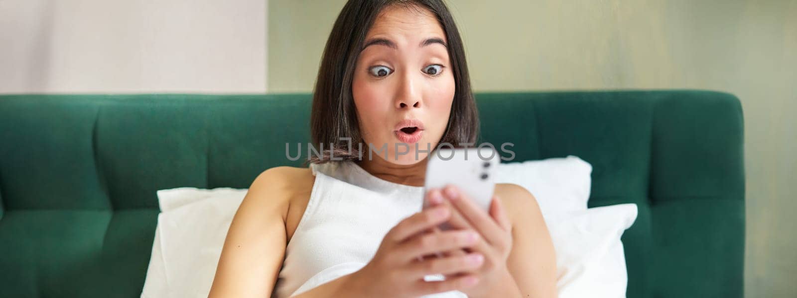 Cute asian girl holding smartphone, looking surprised at mobile phone screen, reading message with amazed face expression by Benzoix