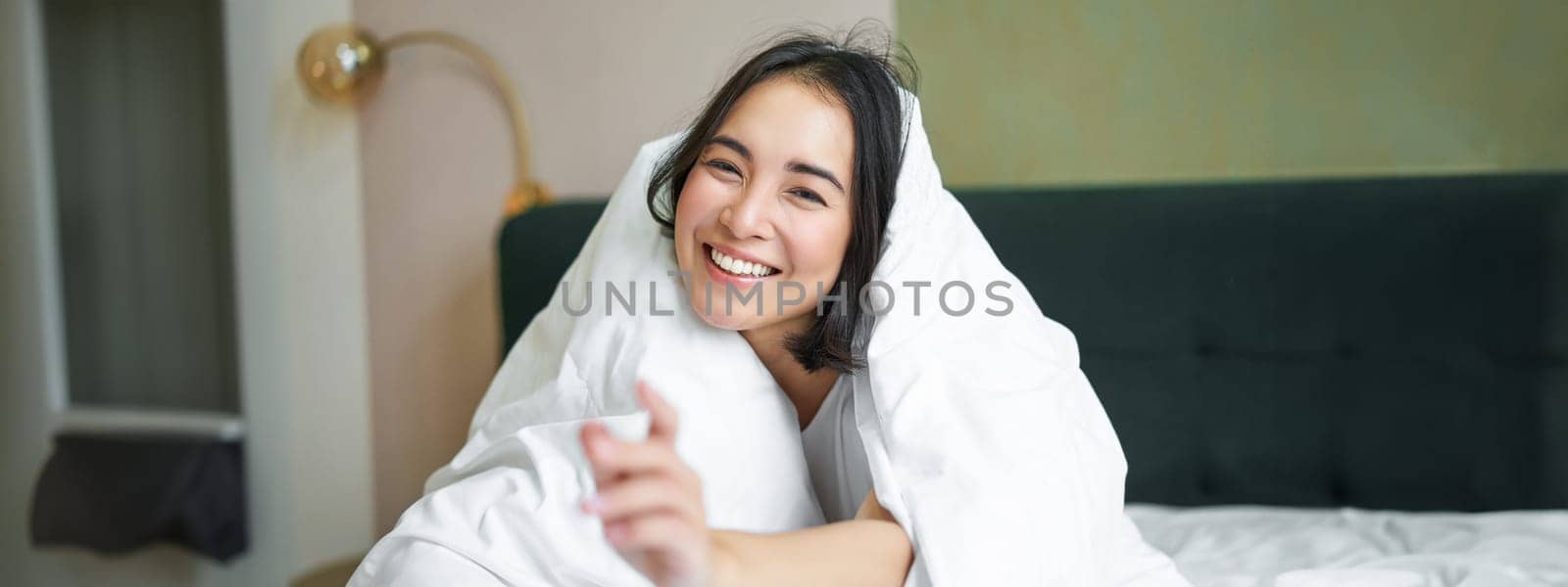 Beautiful asian woman sitting on bed, covered with white duvet, smiling, enjoying happy weekend morning, laughing at camera.