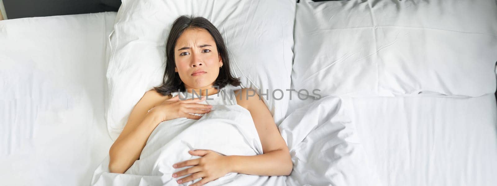 Portrait of sad young asian woman lying in bed, overthinking before going to sleep, stay late because of insomnia, frowning and looking frustrated.