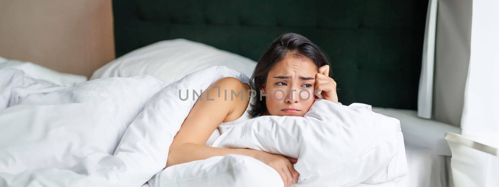 Sad and gloomy asian girl lying in her bed, grimacing and crying, feeling upset, spending time in her bedroom, hugging pillow.