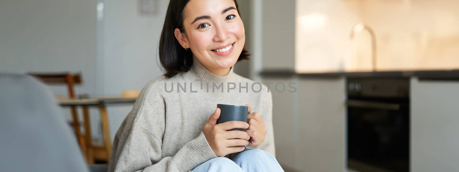Portrait of smiling asian girl with cup of coffee, drinking hot tea and getting cozy at home. Warming up with beverage.