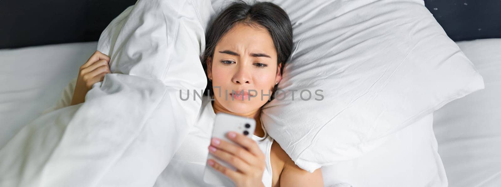 Close up portrait of asian girl lying in bed, looking at smartphone concerned, waking up late and staring at her alarm clock on mobile phone.