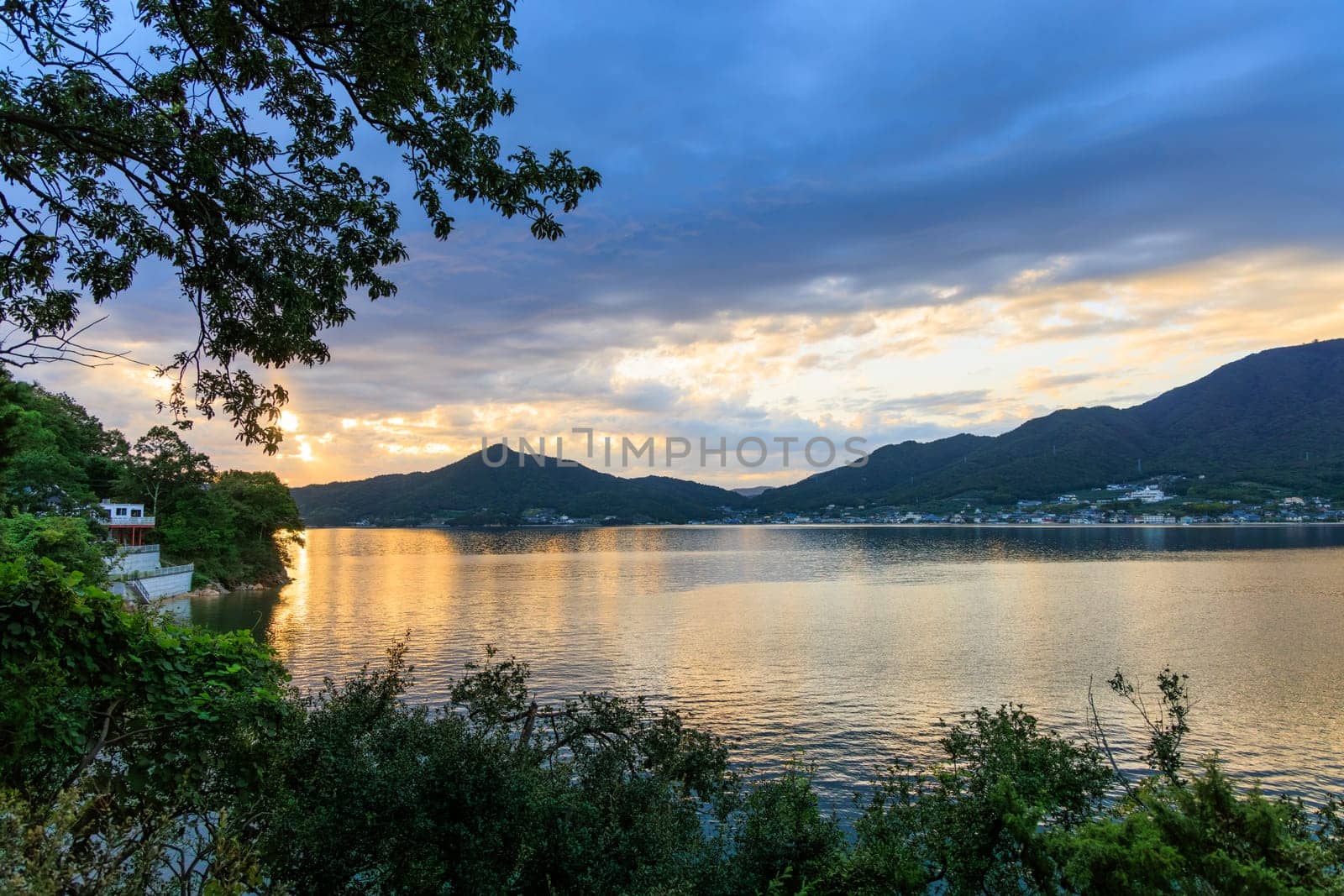 Sunset over calm water in coastal bay by coastal village and mountains by Osaze